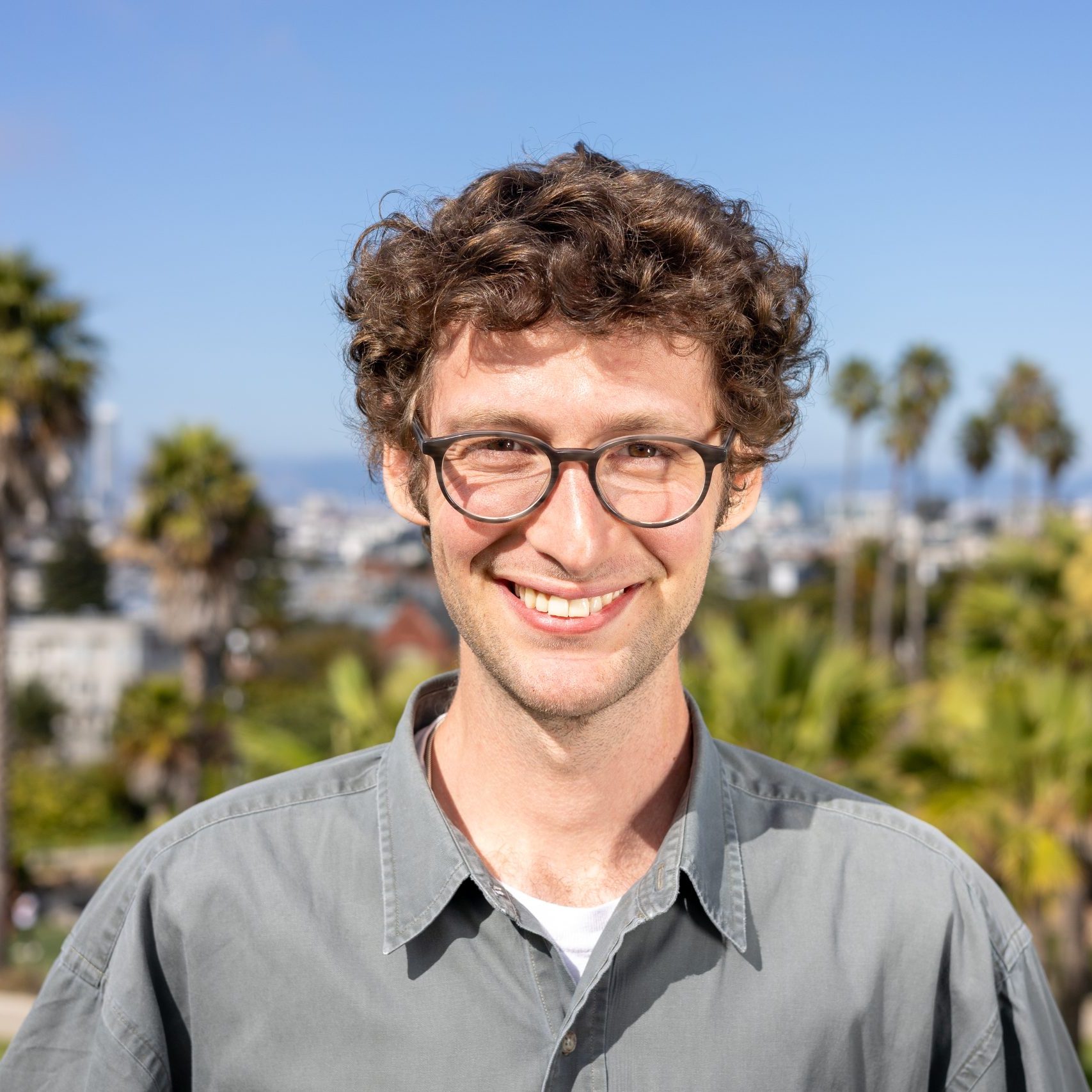 A man with curly brown hair and glasses smiles at the camera, standing outdoors with palm trees and a cityscape in the background.