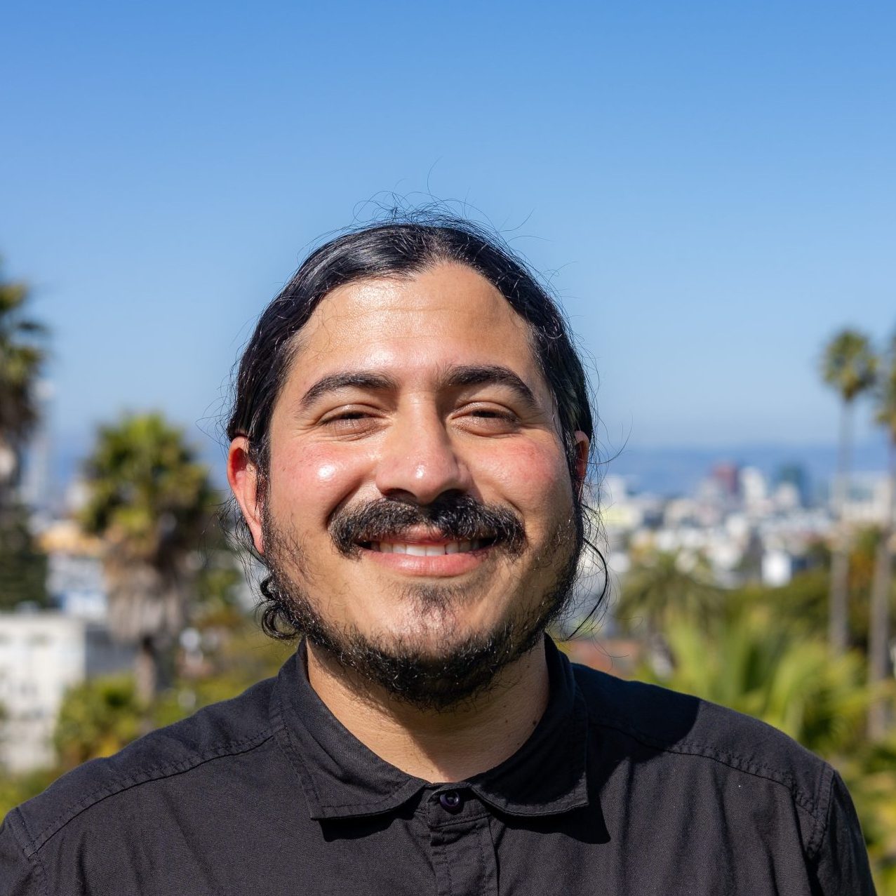 A person with long dark hair and a mustache, wearing a black shirt, smiles outdoors with palm trees and a cityscape blurred in the background under a clear blue sky.