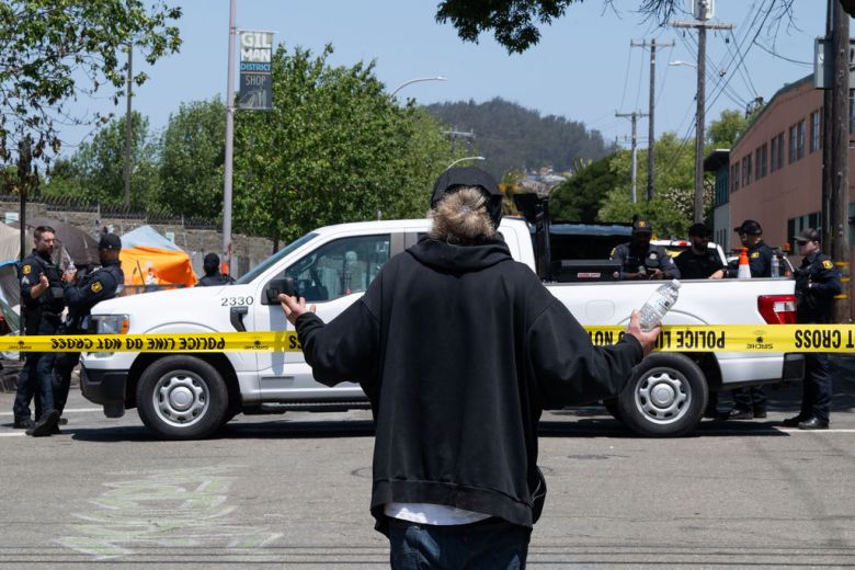 A person stands with their arms outstretched in front of a line of caution tape. Several police officers are on the other side of the line.
