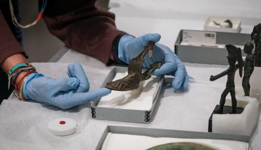 Gloved hands hold a metal piece shaped like a small foot that's believed to have belonged to a piece of furniture. Other bronze figures are on teh table nearby.