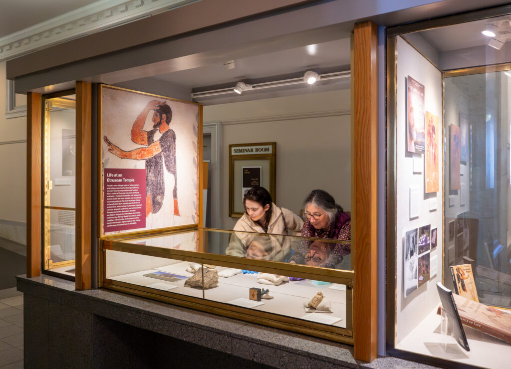 Two people lean over a glass display case, arranging artifacts for an exhibit about the anciet Etruscans at in the Doe Library lobby.