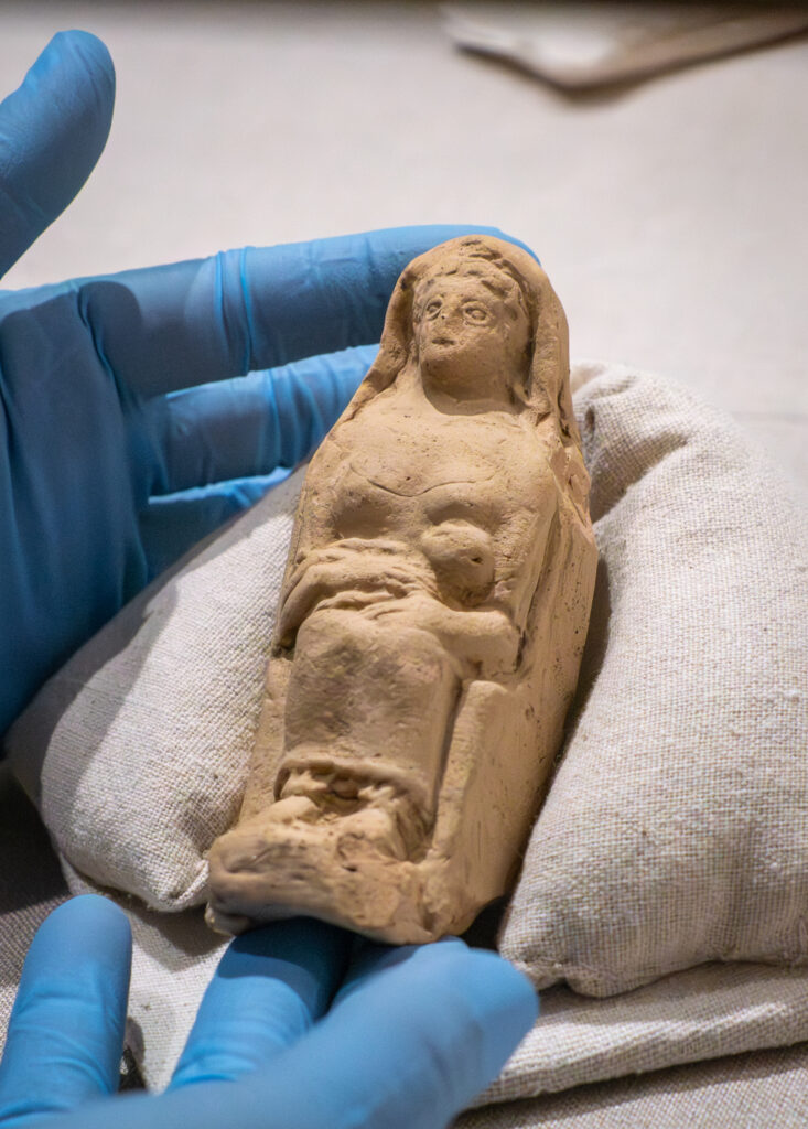 A tan-colored terracotta statuette of seated woman with infant, held in a gloved hands. The object is on display in "The Etruscans Uncovered" exhibit in the Brown Gallery of Doe Library.