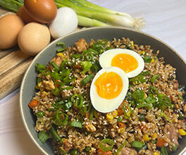 Any grain chicken fried rice is served in a gray bowl with scallions and fresh eggs on a bamboo cutting board in the background.