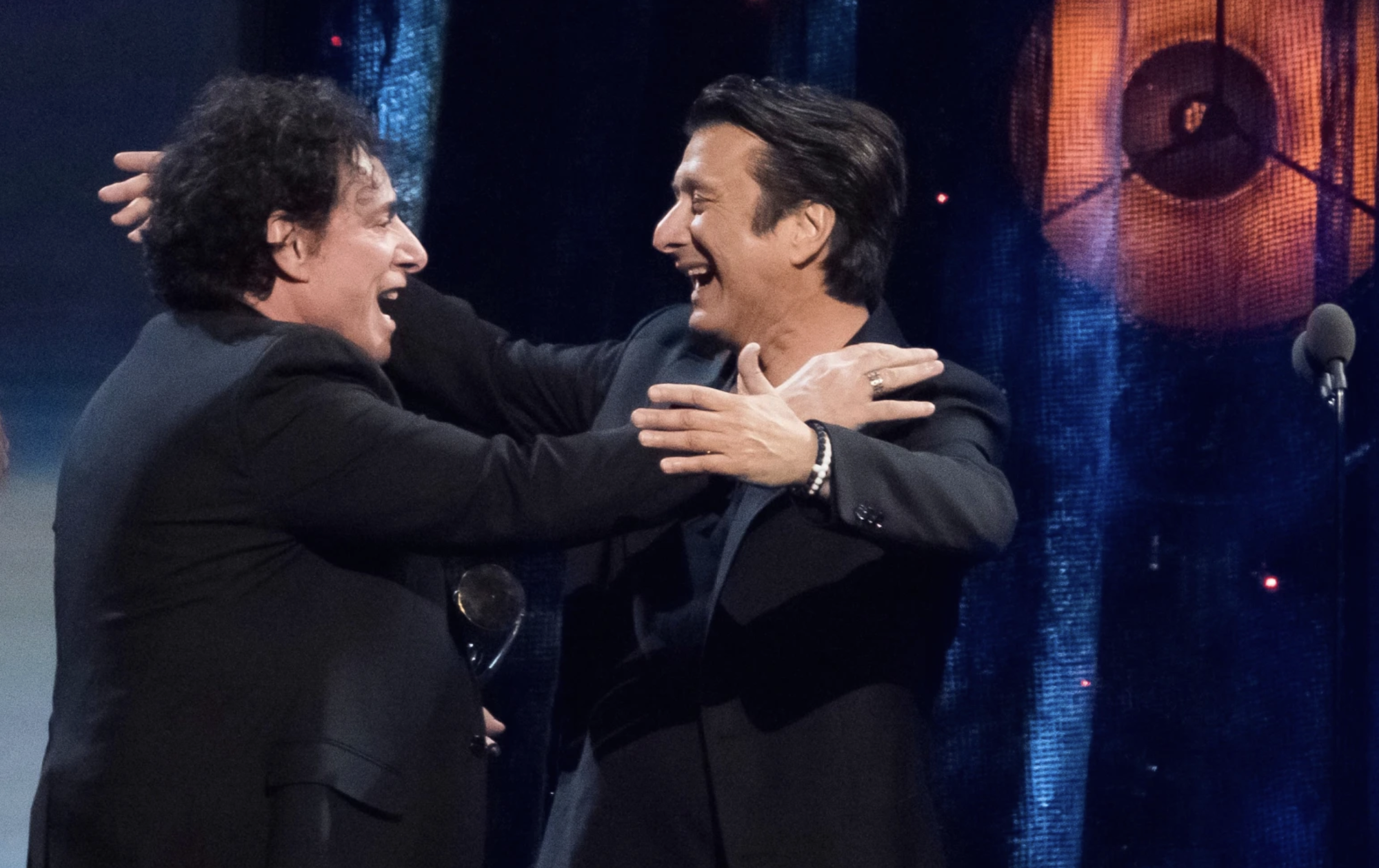 Two white men in black suits smile at each other and begin to embrace, on a dimly lit stage.