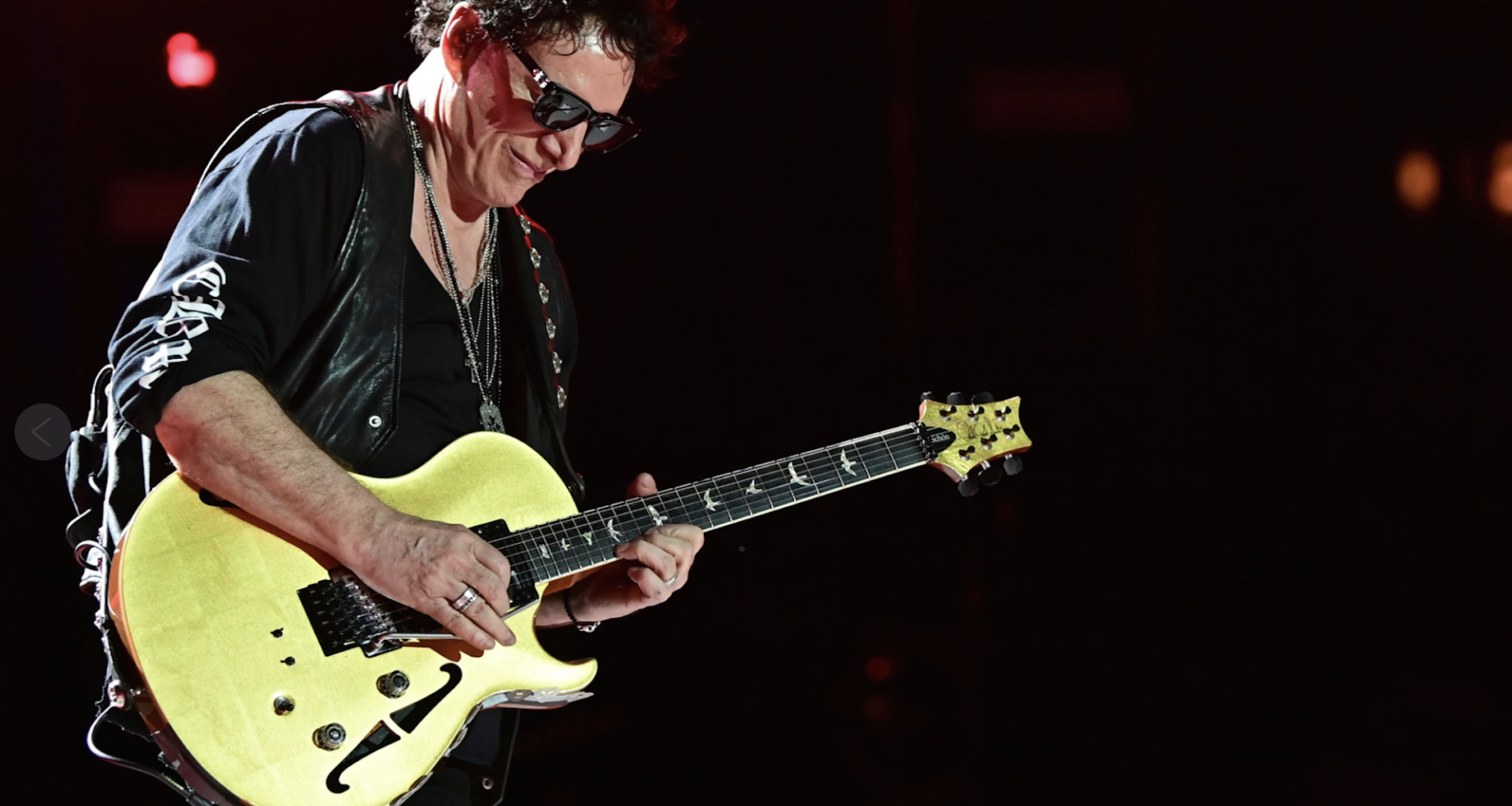 A senior white man with brown hair, dressed in all black and sunglasses plays a yellow electric guitar on stage.