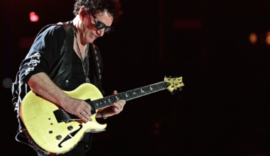 A senior white man with brown hair, dressed in all black and sunglasses plays a yellow electric guitar on stage.