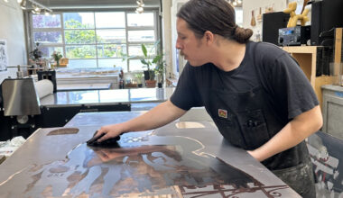 A printmaker in a black apron wipes a large metal printing plate on a worktable in a bright printmaking studio.