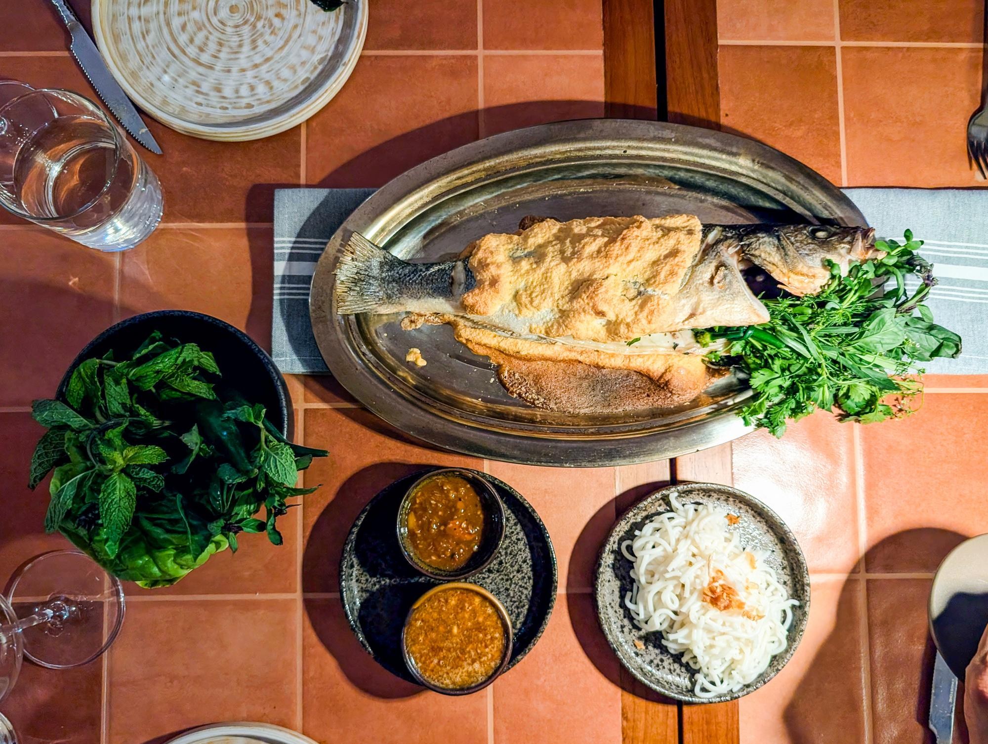 A platter of whole fish partially in salt crust, with herbs sticking out, next to a plate of dishes, a plate with two sauces, and bowl of mint, at Saam in San Francisco.