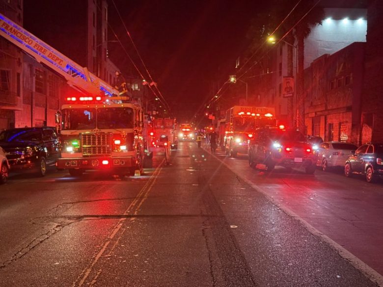 Multiple fire trucks and emergency vehicles with flashing lights are parked on both sides of a city street at night.
