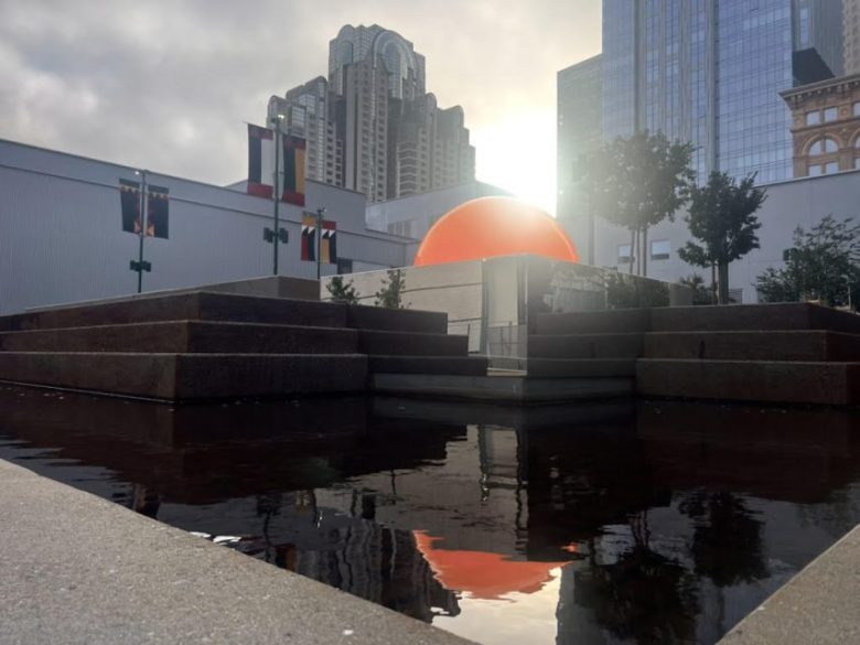 Modern cityscape with buildings, flags, and a large orange dome, reflected in a still water pool in the foreground at sunset.