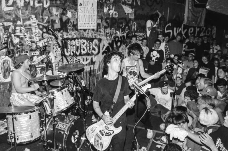 A black and white photo of a young Green Day performing in front of a packed audience. A graffiti-covered wall is behind them on a small stage