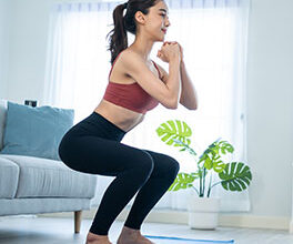 A dark-haired young woman with a ponytail does squatting exercises on a blue mat.