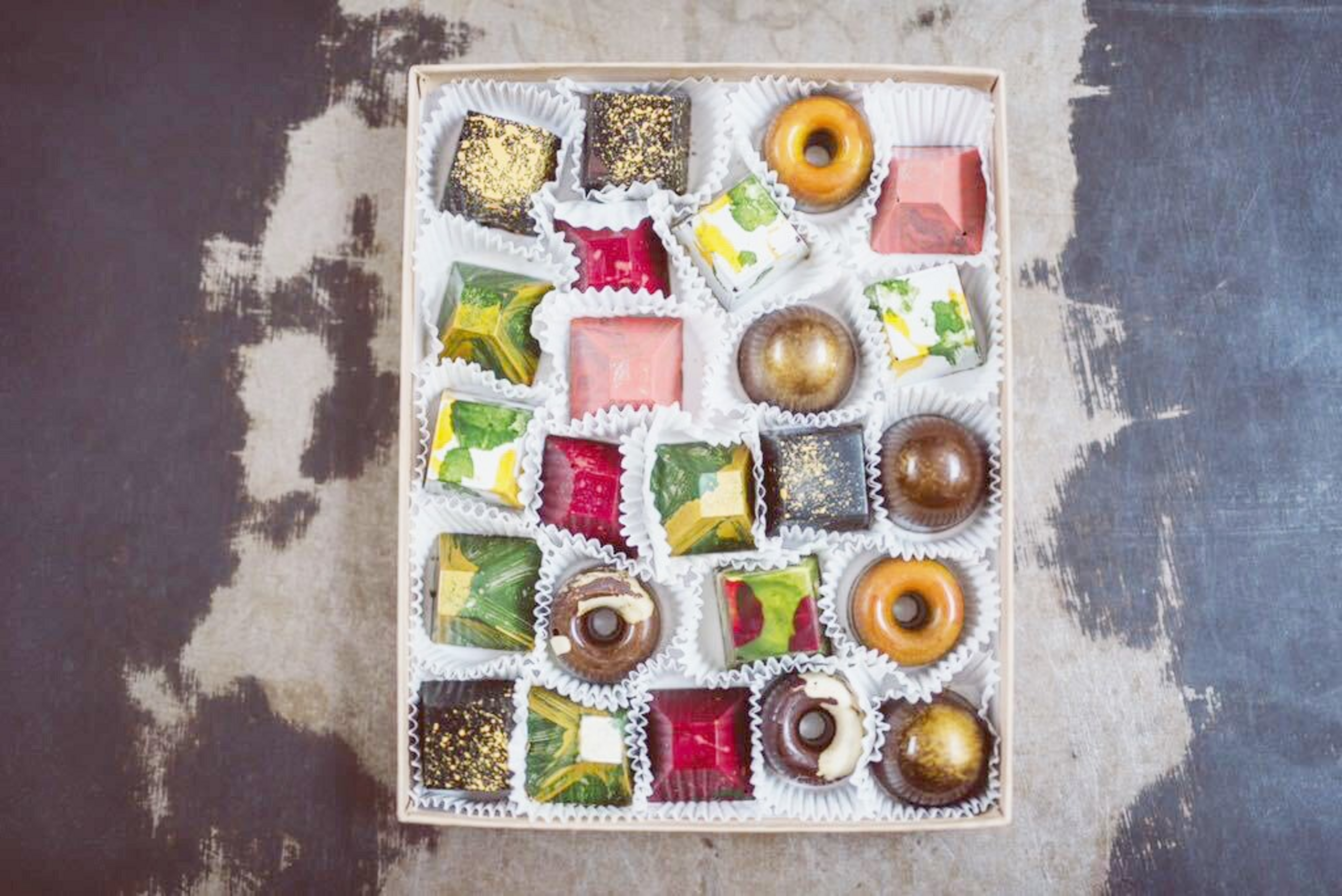 A box contains an assortment of colorful, artistically designed chocolates in various shapes like squares, domes, rings, and rectangles, each in paper cups.