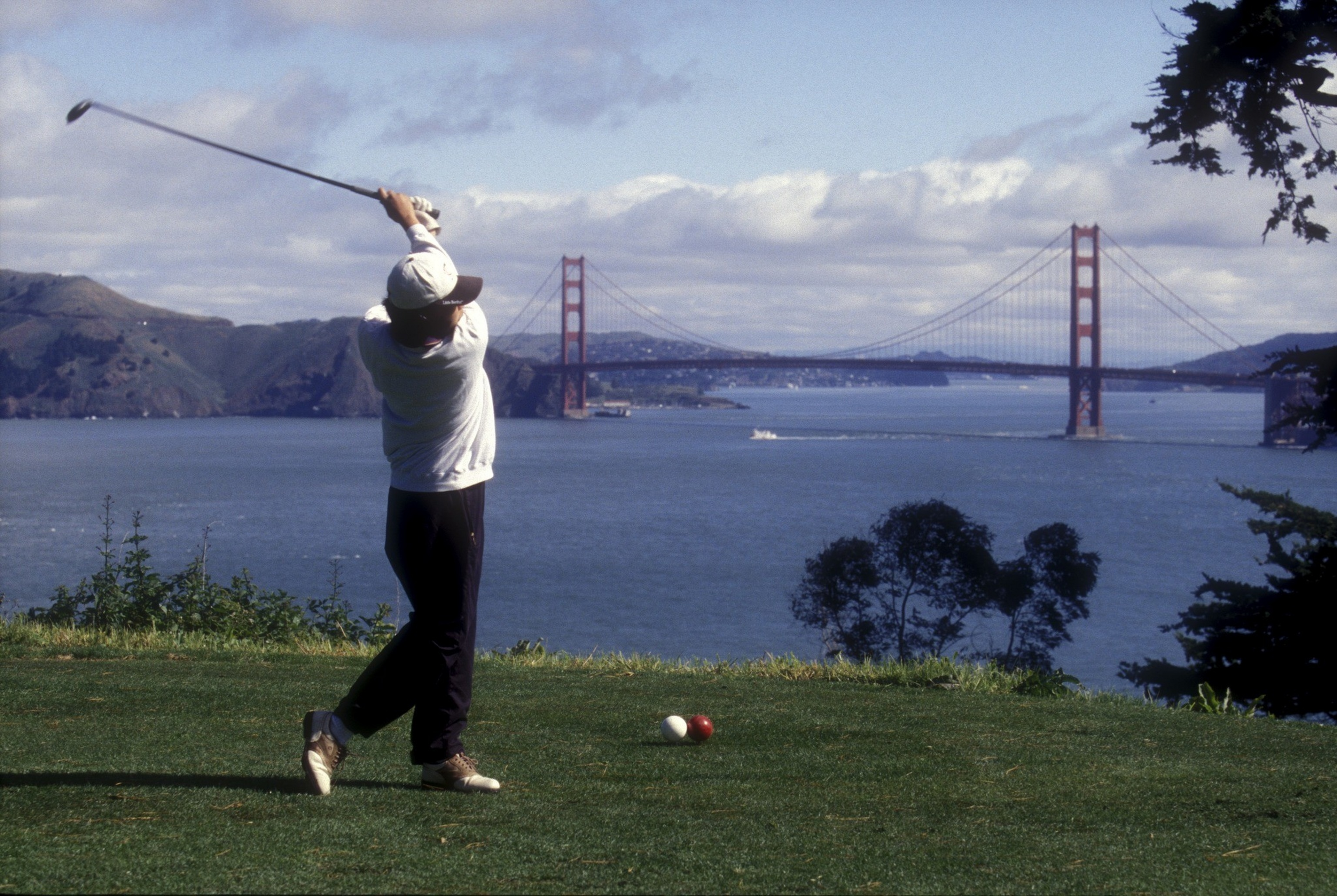 A golfer in mid-swing stands on a grassy area overlooking the San Francisco Bay, with the Golden Gate Bridge and hills in the background.