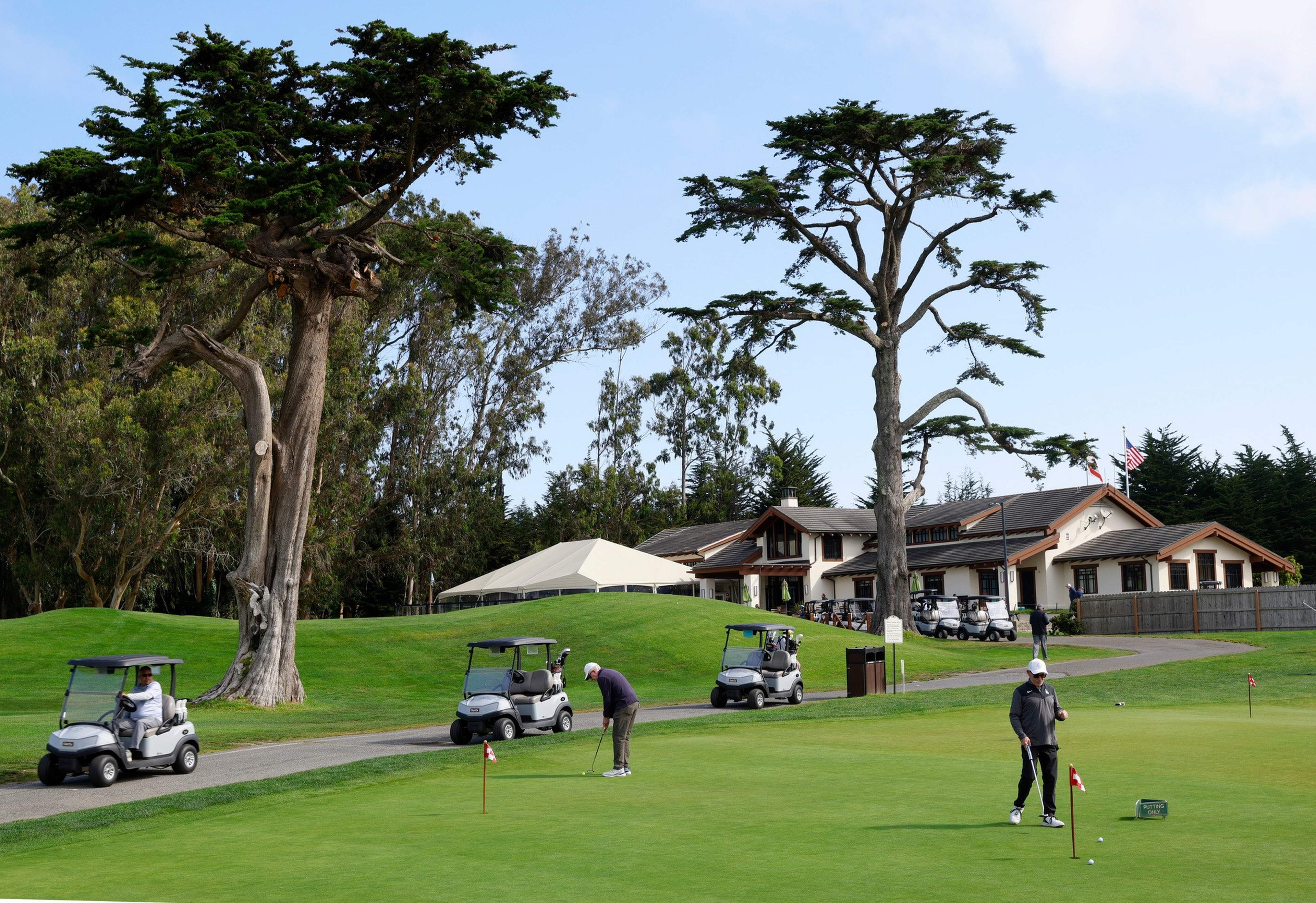 Two people are playing golf on a green near a clubhouse with several golf carts parked nearby and tall trees in the background.
