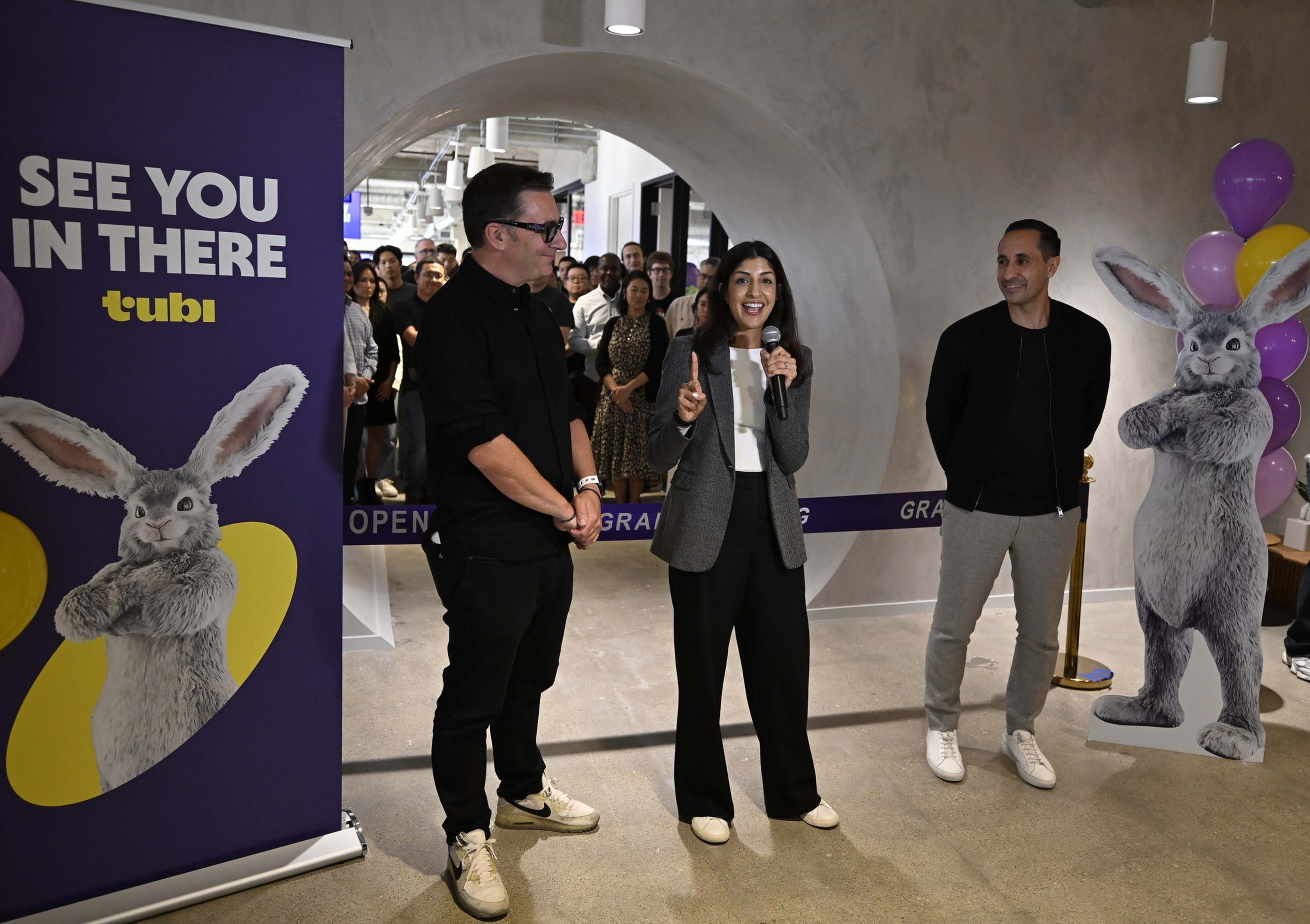 Three people stand in a modern hallway, one woman speaking with a microphone, with a crowd behind and two large rabbit cutouts on either side.