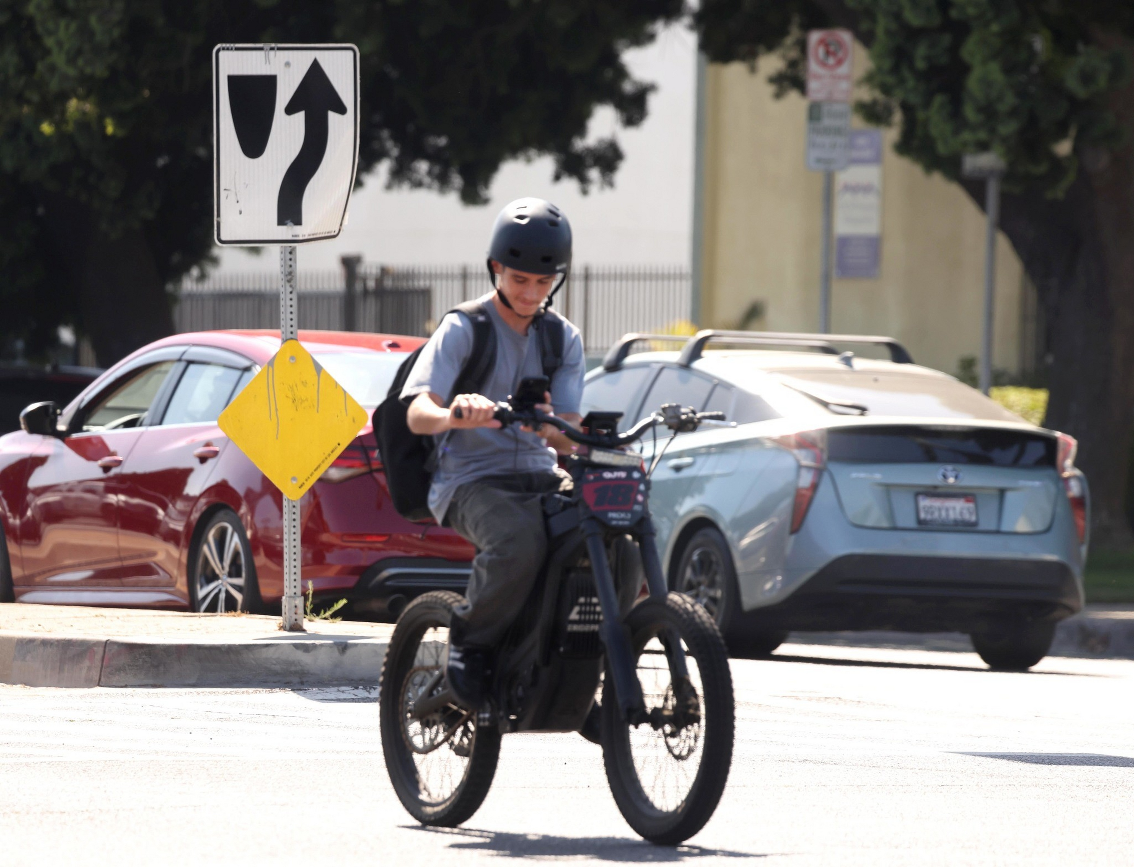 A person wearing a black helmet rides an electric bike on a street near a traffic sign, with two parked cars and trees in the background.
