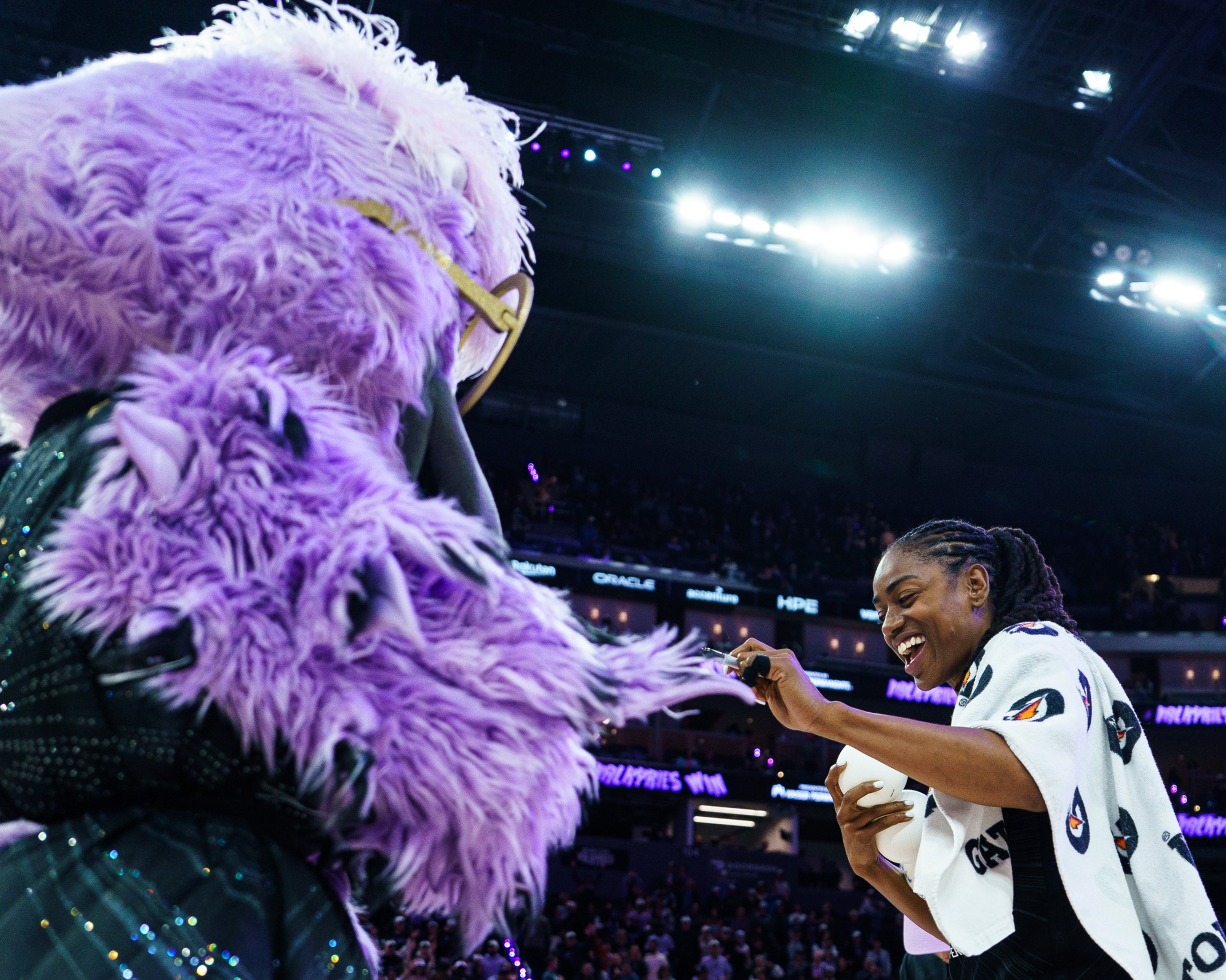 A basketball player smiling and reaching out toward a large purple furry mascot in an arena filled with spectators and bright lights.