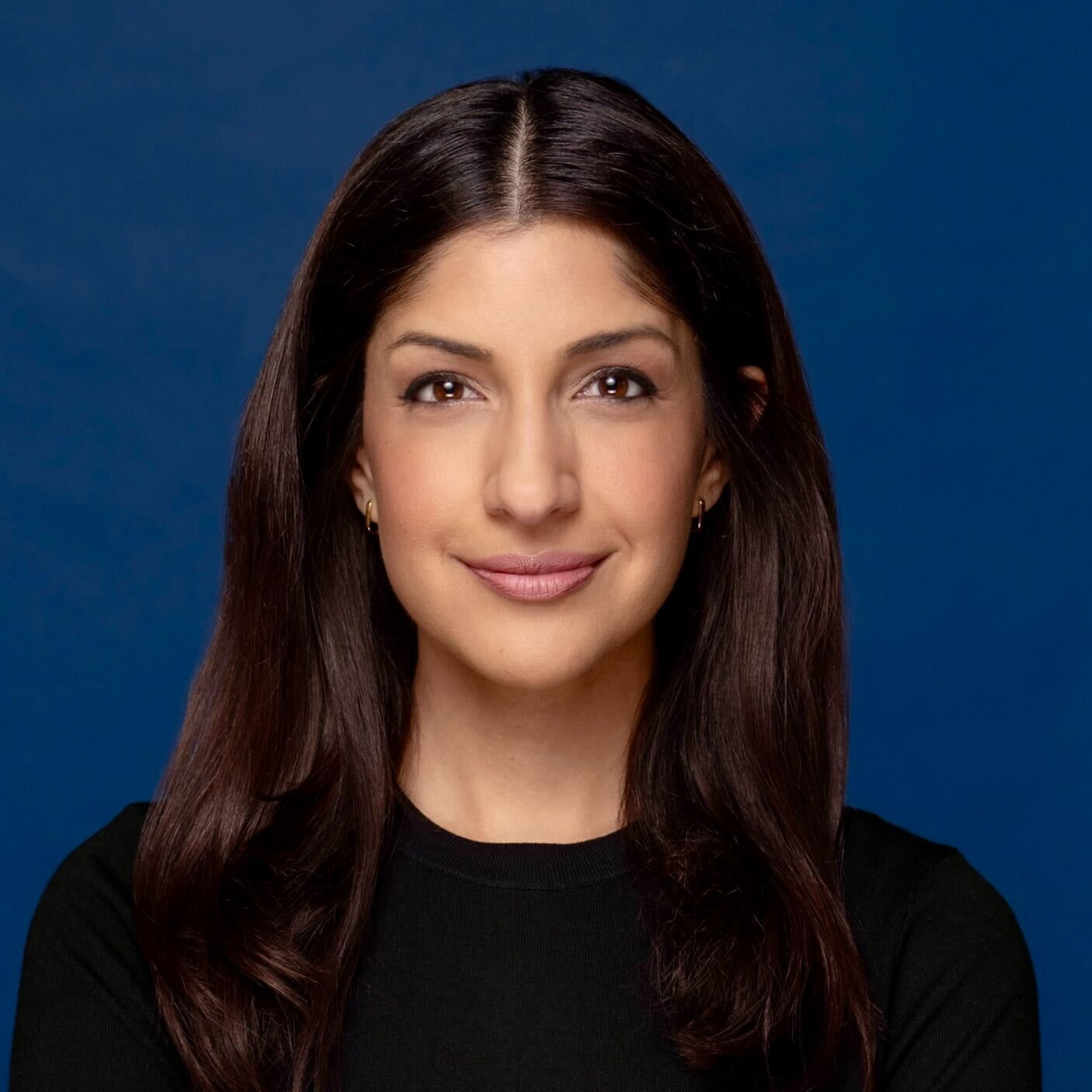 A woman with long, dark brown hair and light brown eyes smiles gently against a solid blue background, wearing a simple black top and small gold earrings.