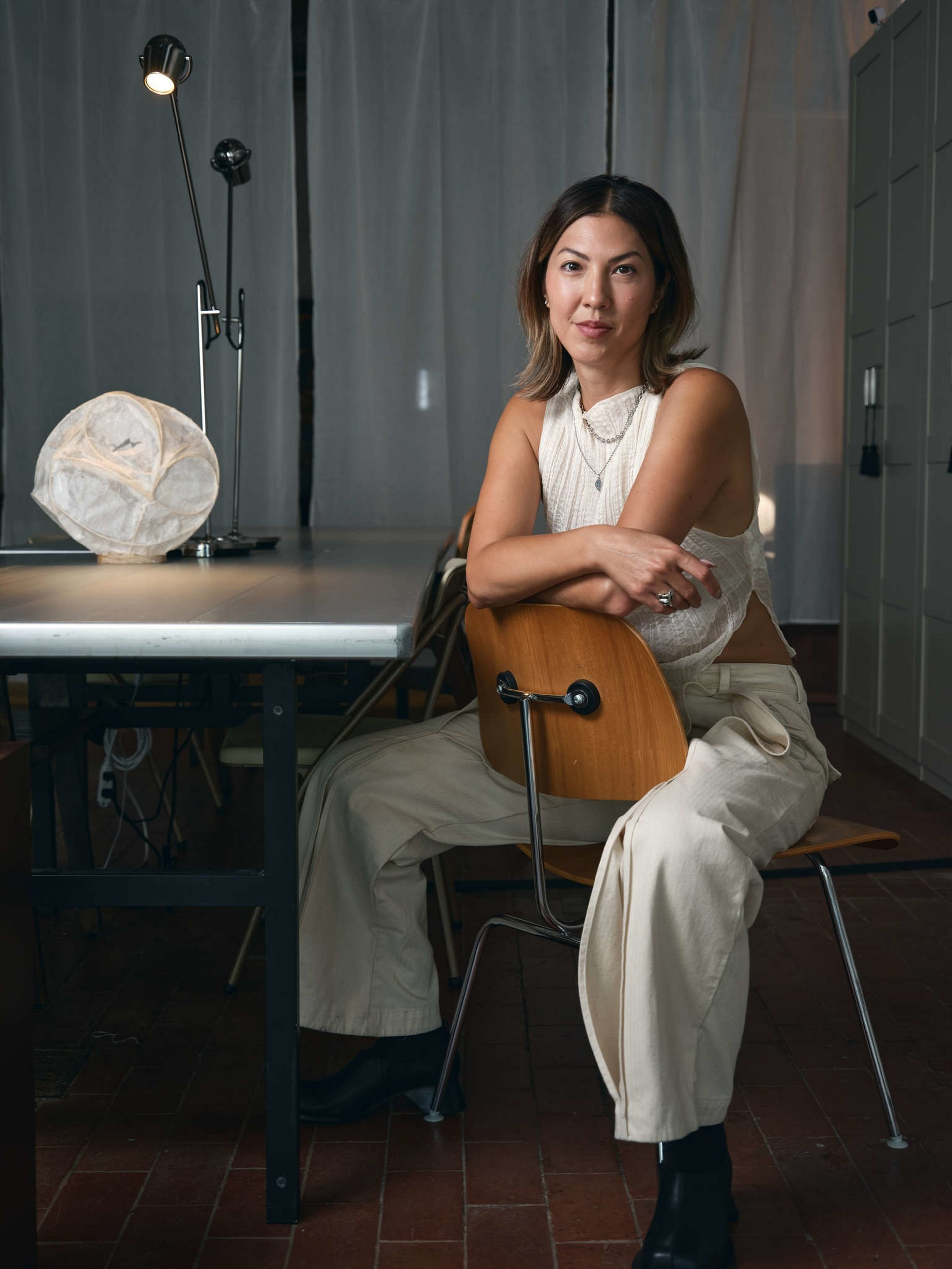 A woman with shoulder-length hair sits sideways on a wooden chair, wearing a sleeveless white top, cream pants, and black boots in a dimly lit room.