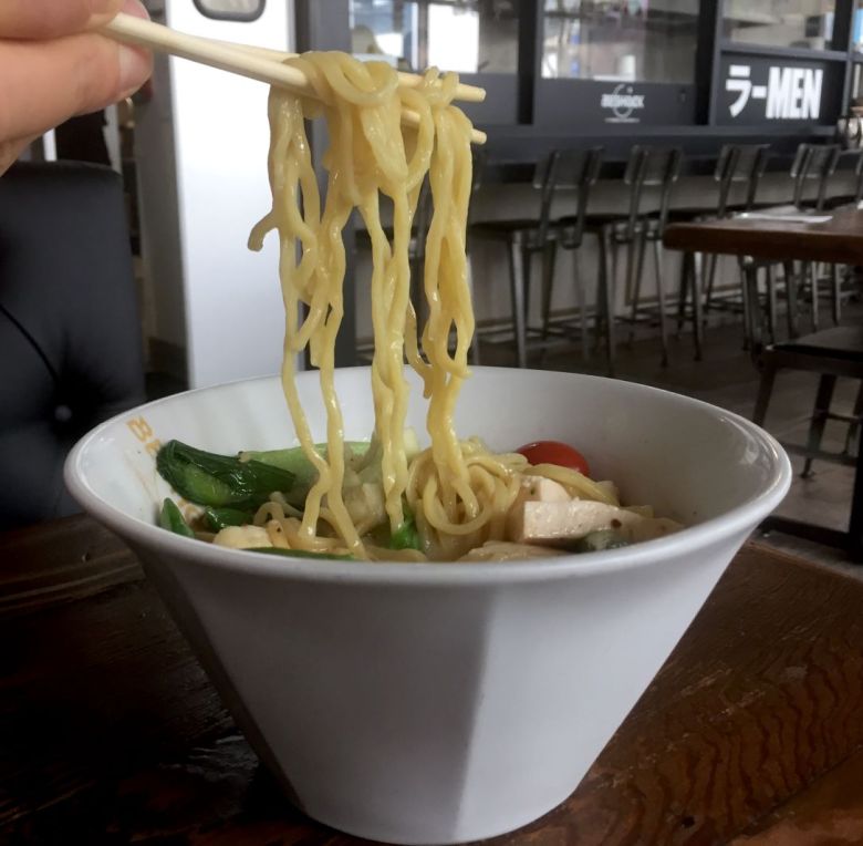 chopsticks are  held over a bowl of ramen, with noodles in the tines. Chairs lined up along a dining bar can be seen in the background.
