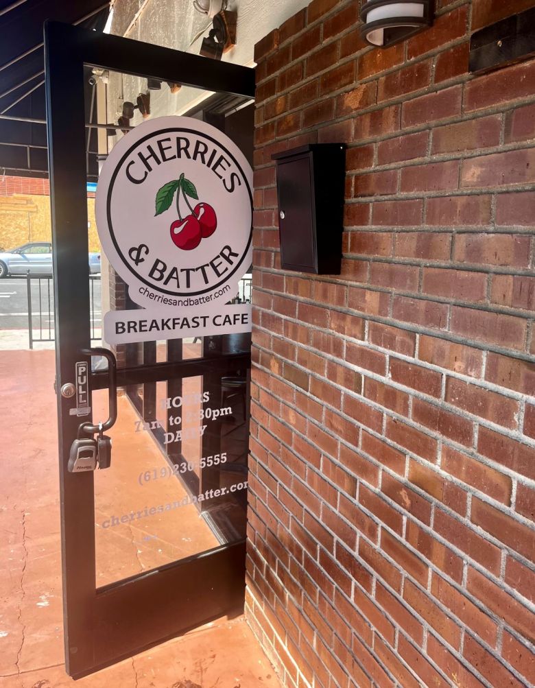 The painted sign for an eatery, on the glass entry door, set in a brick facade. It reads "Cherries & Batter. Breakfast Cafe" 