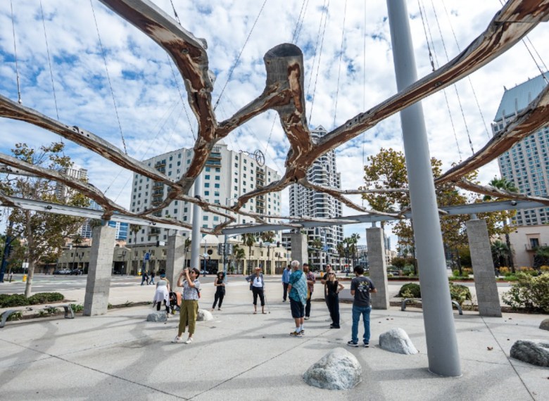 A group of people tour an outdoor plaza beneath large metal beams that are straight or resemble tree limbs.