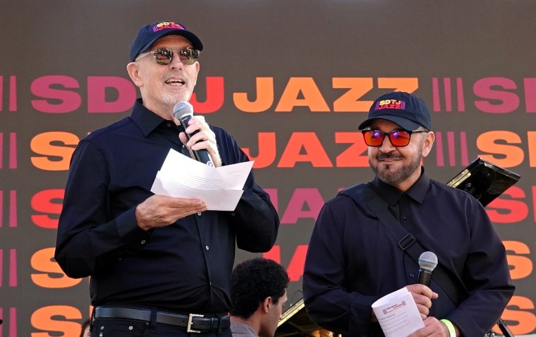 Two men stand on a stage, with the one on the left speaking into a microphone. The word "jazz" can be seen repeated on the backdrop behind them.