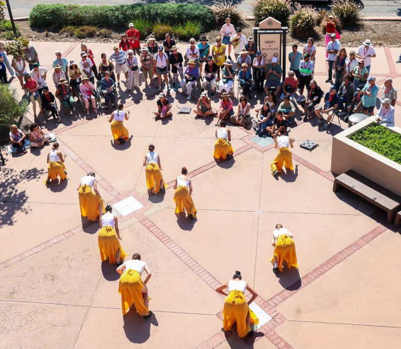 Two people stand at the top of an outdoor plaza, with four people lined up behind them and five more in the back. Spectators line two sides of the plaza, watching.