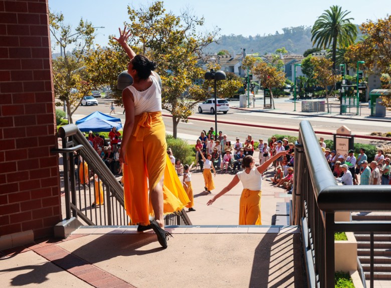 A woman reaches to the sky as she starts to head downstairs to a plaza, where dancers dressed like her, in while tops and orange pants, perform for people standing outdoors with a street visible behind them.