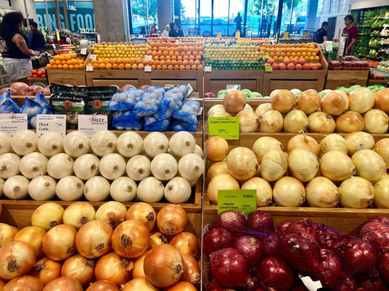 Bins filled with produce, including several varieties of onions sit in front of more produce, including oranges and lemons, in a supermarket.
