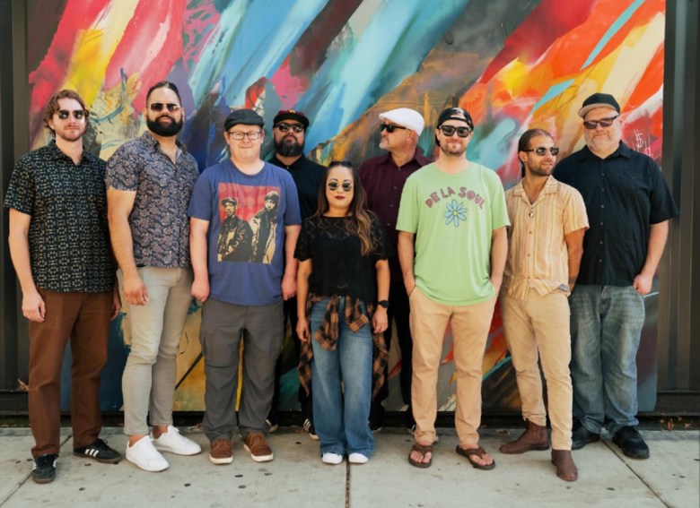 Eight men, and a women in the center, all dressed casually, stand in front of a colorful wall.