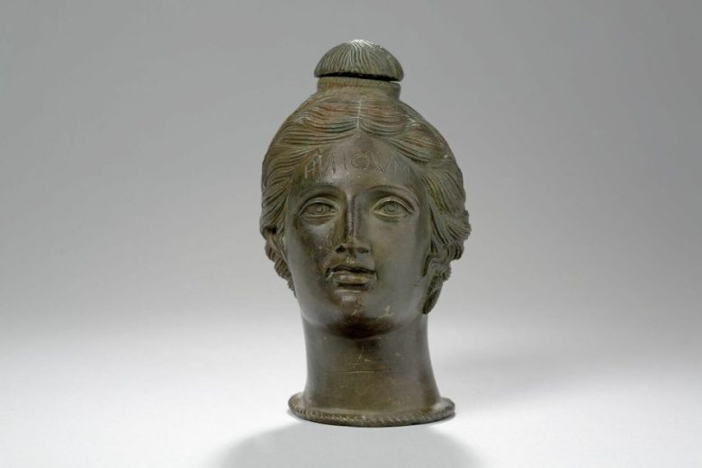 Bronze vessel shaped like a human head, with detailed facial features and a rounded lid on top, set against a plain background.