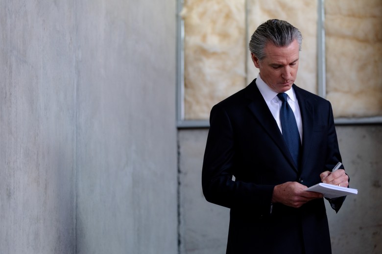A lawmaker with slick-back gray hair and wearing a dark suit, looks down as they write on a small white book.