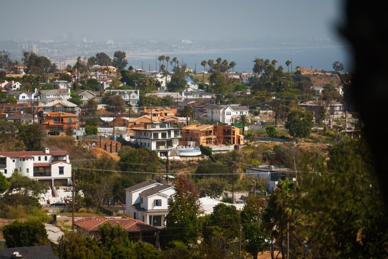 A hillside neighborhood of modern homes, trees, and winding streets overlooks a calm blue sea beneath a hazy sky, creating a quiet coastal atmosphere.
