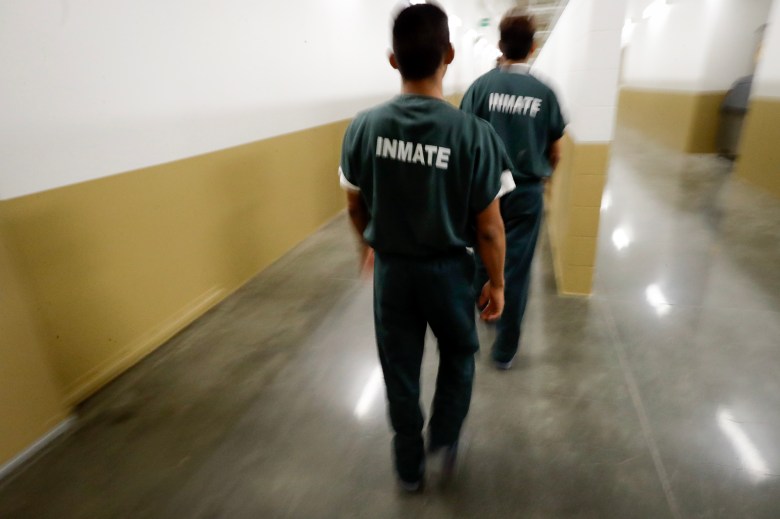 Two people wearing uniforms labeled “INMATE” walk down a hallway inside a detention facility.