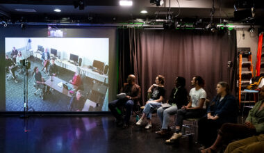 Students seated in a row of stools face a large projection screen showing a video call with collaborators, inside a darkened studio space with theater lighting rigs overhead.