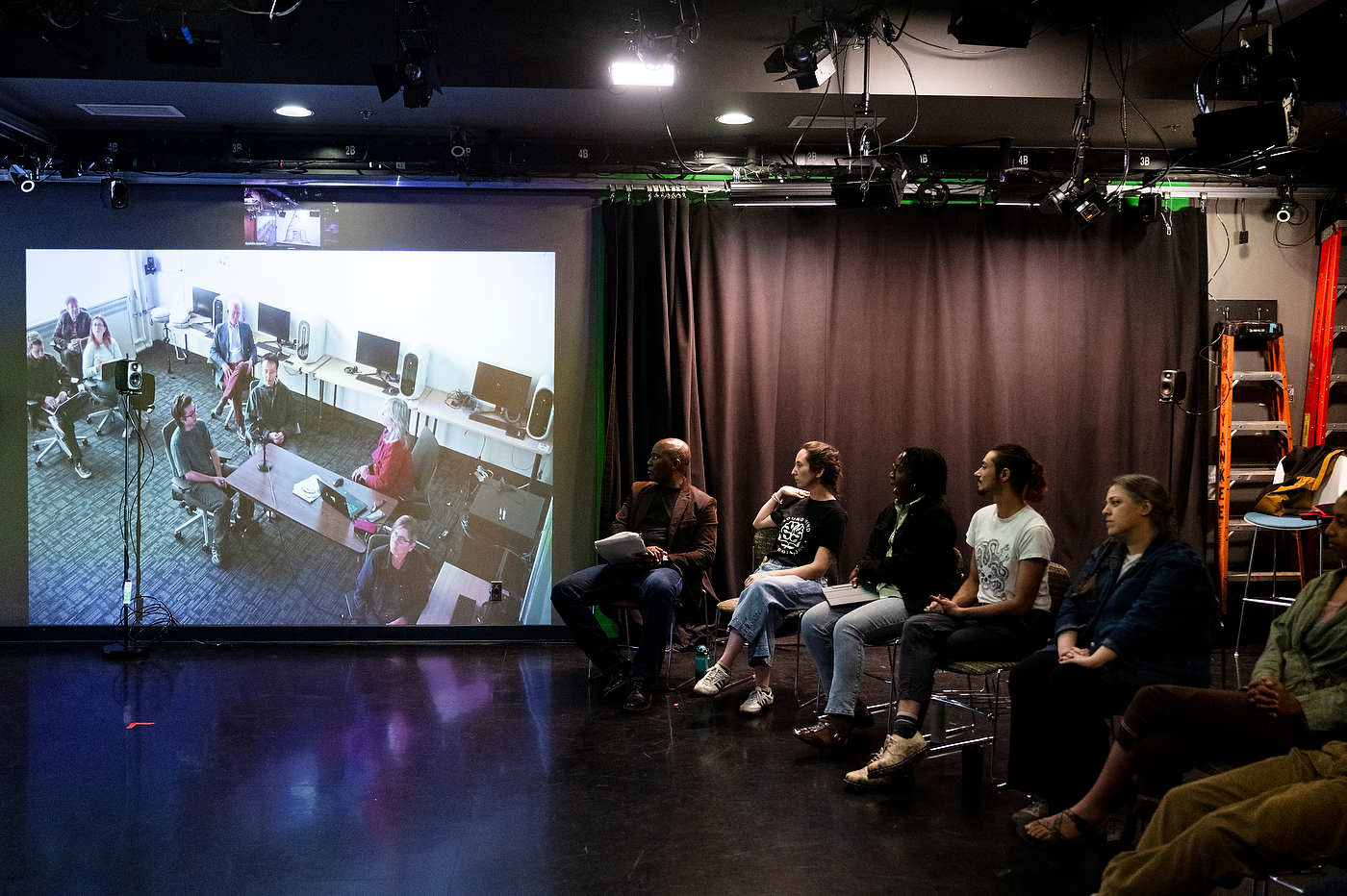 Students seated in a row of stools face a large projection screen showing a video call with collaborators, inside a darkened studio space with theater lighting rigs overhead.