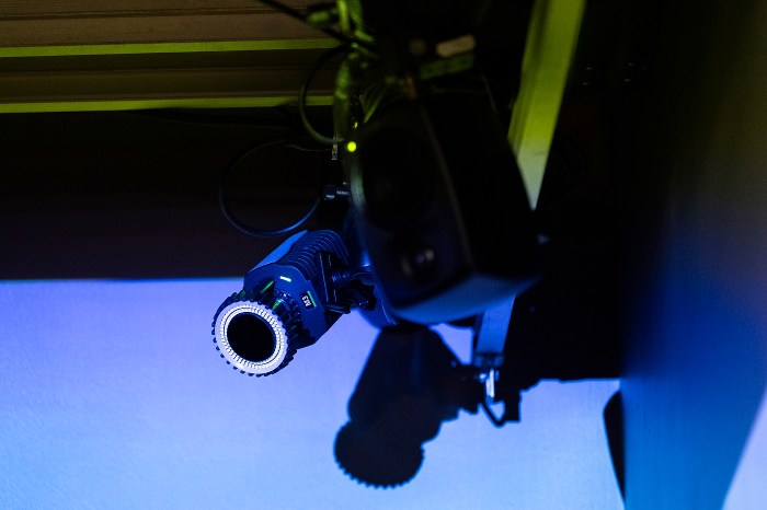A studio camera or projector mounted on rigging, lit in blue light.