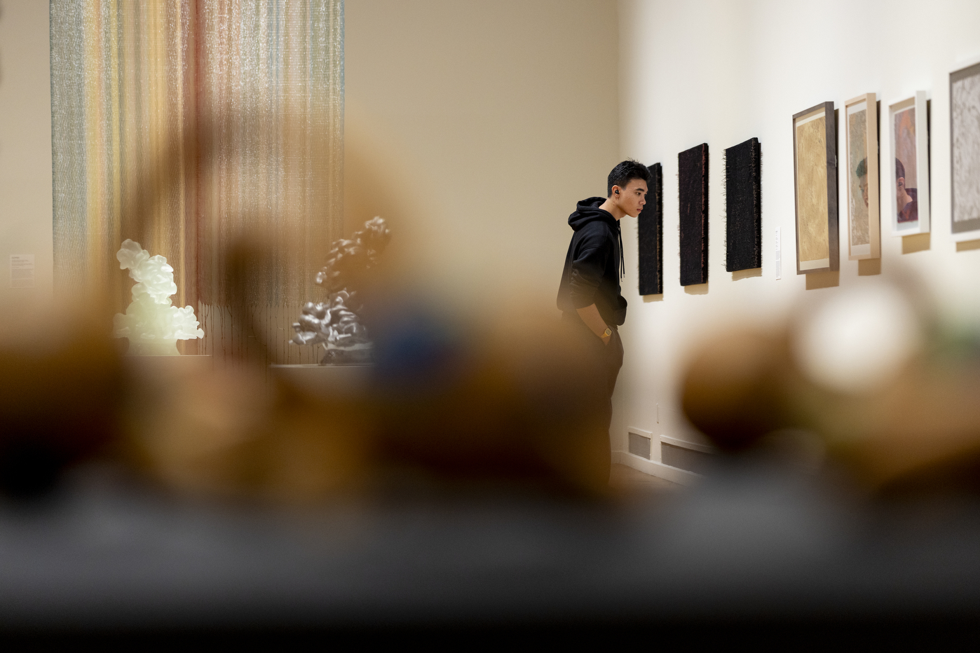 A visitor views a row of textile and framed artworks on a gallery wall, with sculptures blurred in the foreground.