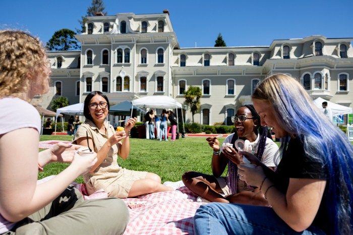 Students seated on a picnic blanket eating and laughing, with a campus building in the background.