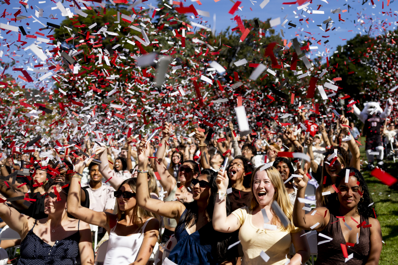 Students cheering with arms raised as red, white, and black confetti fills the air above the crowd.