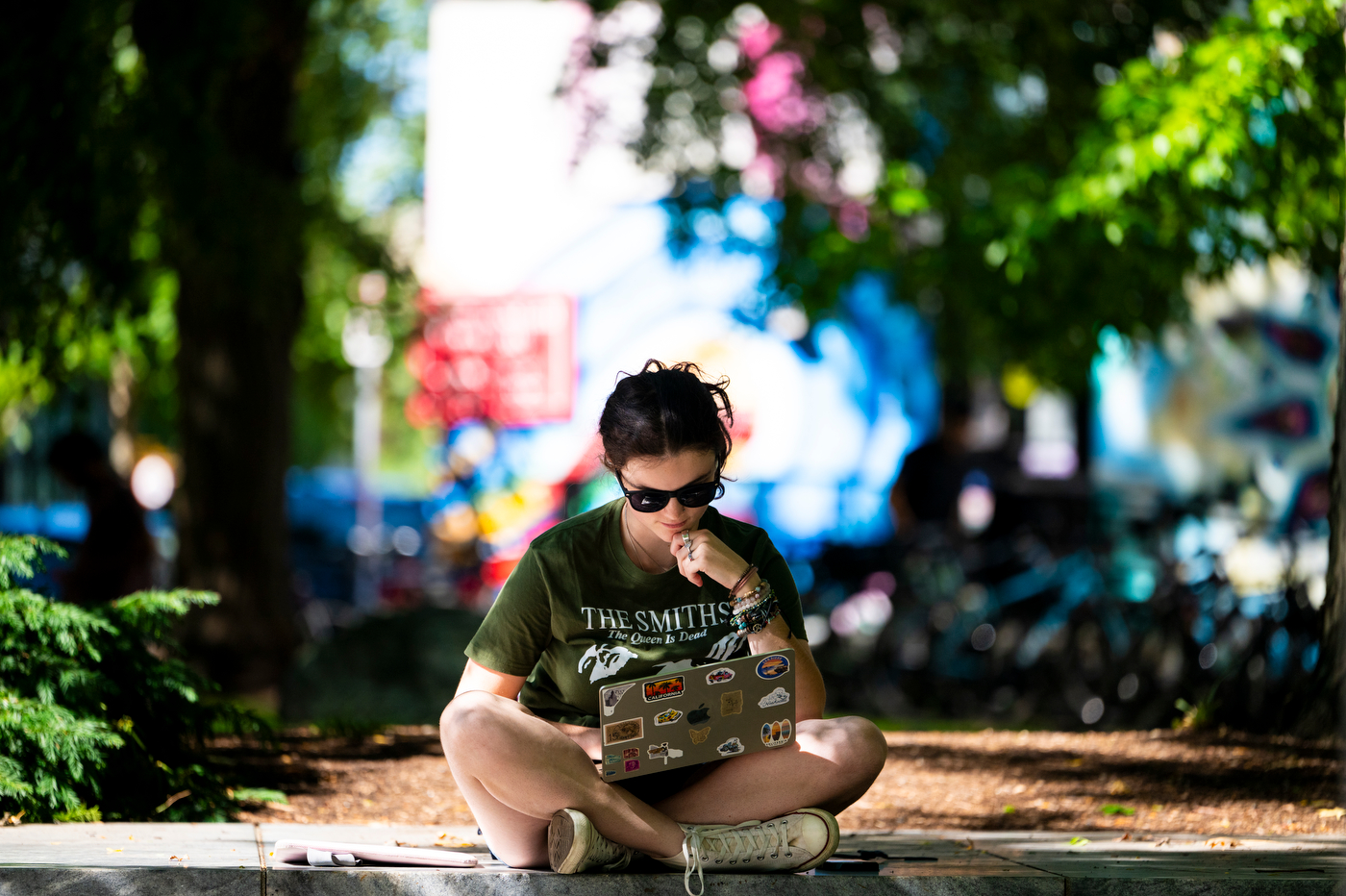 A student sits on the ground studying with a laptop near the Veterans Memorial, surrounded by trees and murals.