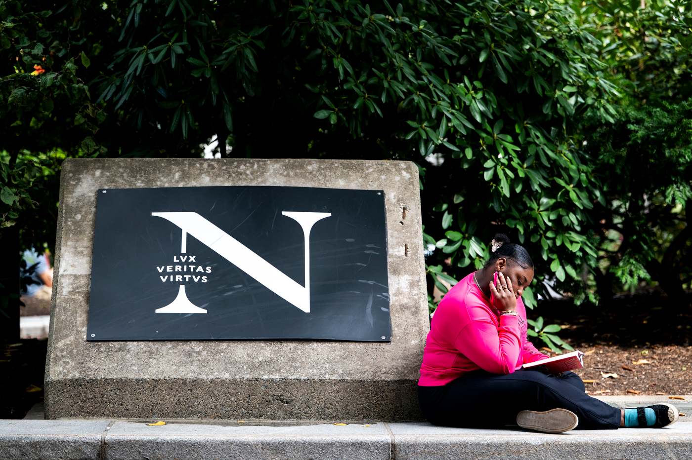 Habigai St Jean reads a book beside a stone Northeastern University "N" sign in Snell Quad.