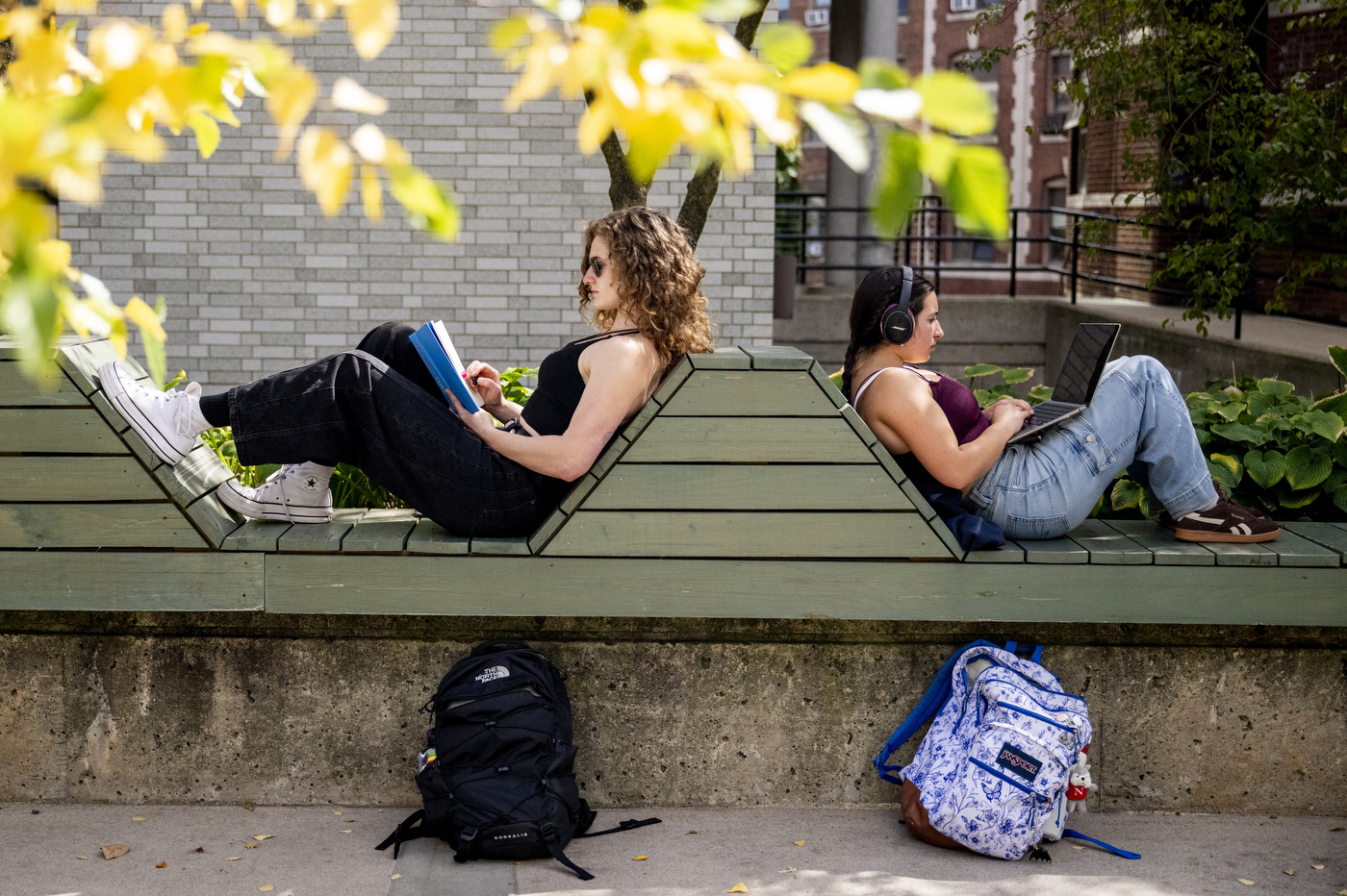 Two students recline on a zigzag-shaped bench outdoors, one reading a book and the other using a laptop.