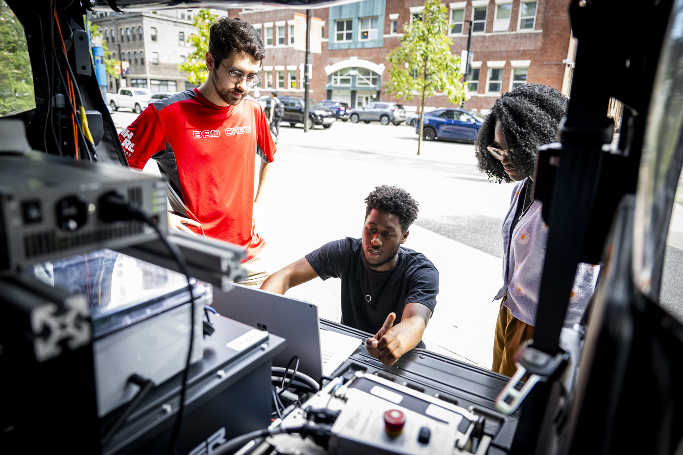 Aidan Reichenberg, James Tukpah, and Wisena Joseph examine equipment on an autonomous vehicle parked on a city street.