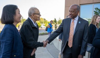 Elderly man with cane shakes hands, smiling with a suited official outdoors
