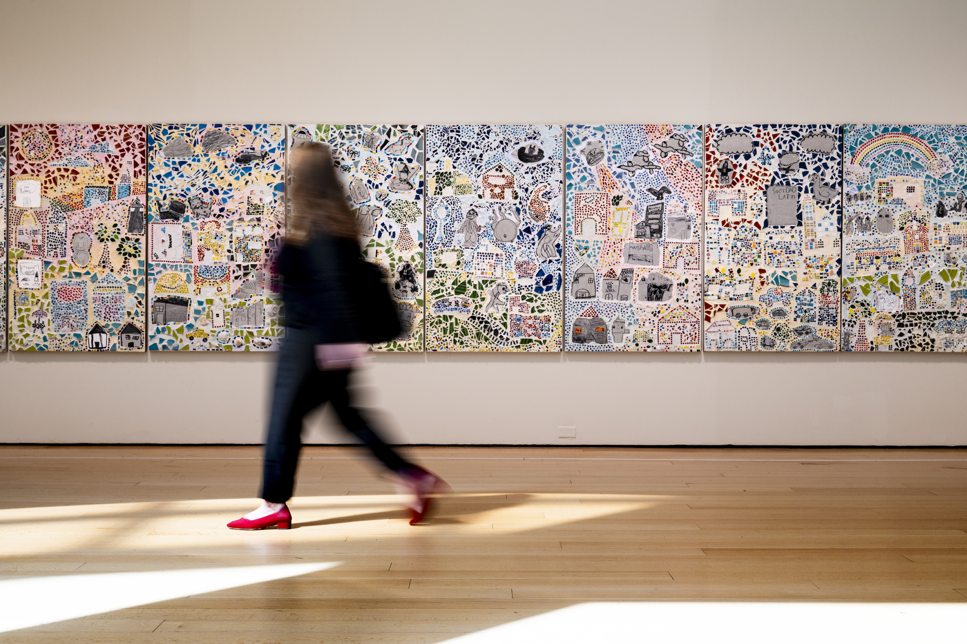 A museum visitor walks past a series of mosaic art panels lining a gallery wall.