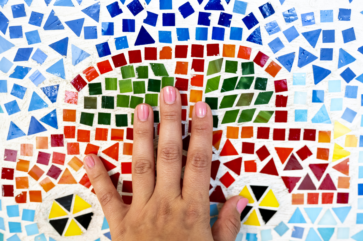 A hand touches a colorful mosaic artwork featuring a rainbow made of small tiles.