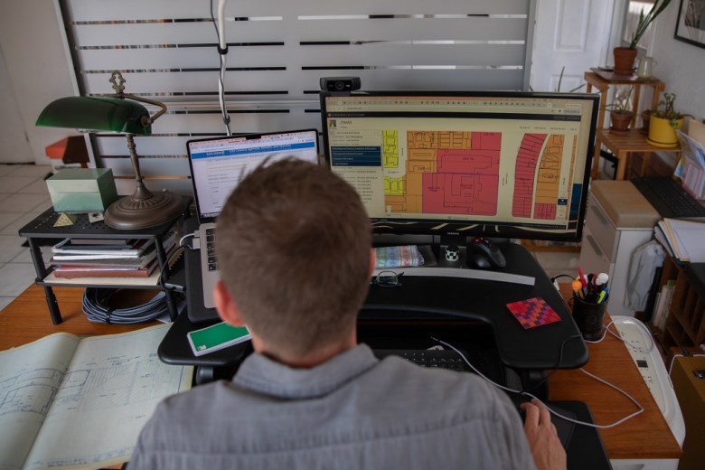 A person sits at a computer workstation, viewing a colorful floor plan on a large monitor; the tidy office setting feels focused and professional.